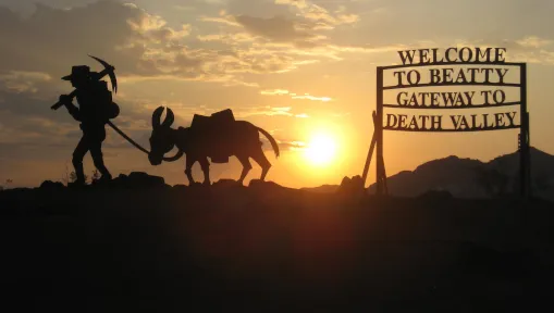 Silhouette of a prospector and burro beside the Welcome to Beatty, Gateway to Death Valley sign at sunset
